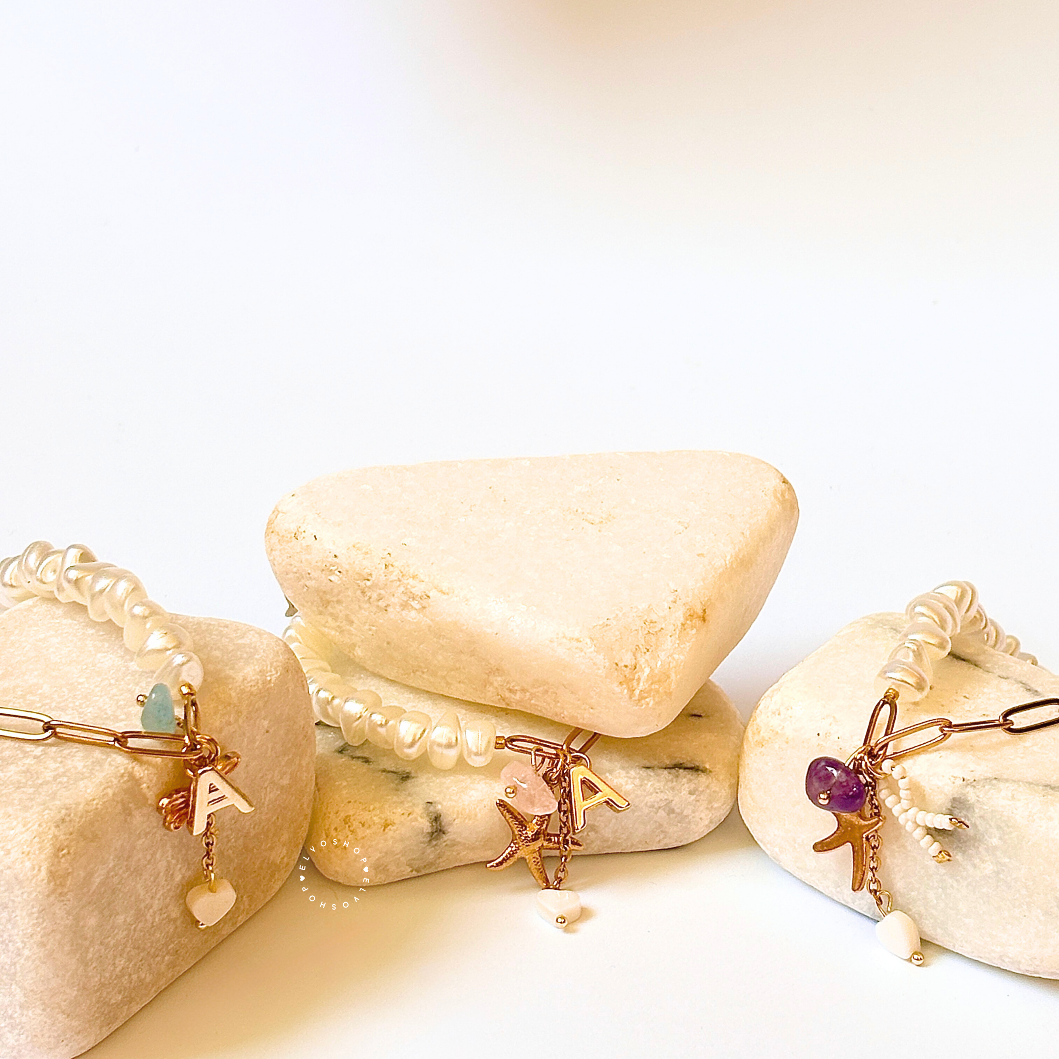 Three pearl beaded bracelets with gold-toned charms (letter 'A', starfish, and heart) displayed on stacked natural stones against a white background.