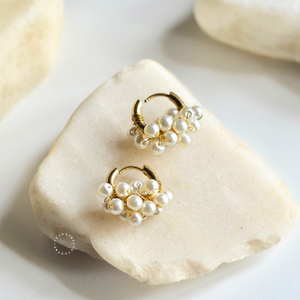 A pair of gold huggie hoop earrings adorned with clusters of small white faux pearls, displayed on a light beige stone against a soft white background.