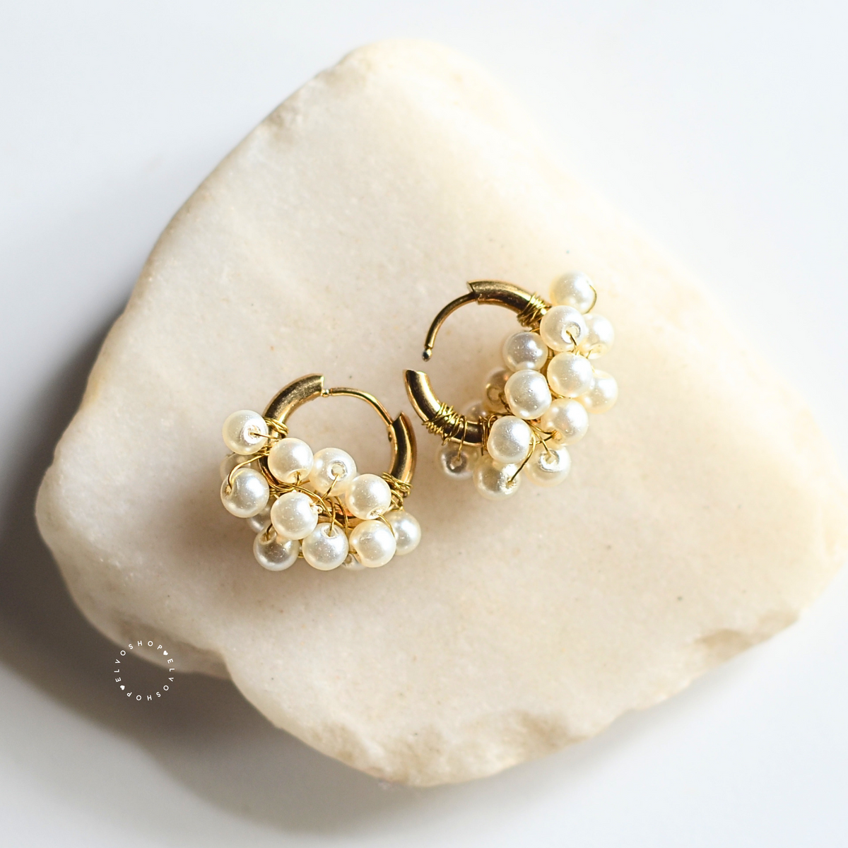 A pair of gold huggie hoop earrings adorned with clusters of small white freshwater pearls, displayed on a light beige stone slab against a clean white background.