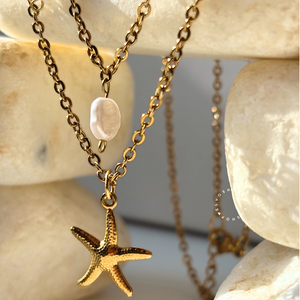 A layered necklace set featuring a textured gold starfish pendant, a white pearl bead, and a gold chain, styled on a background of smooth beige stones.
